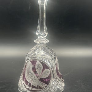 May include: A clear crystal bell with a long handle and a diamond-cut pattern. The bell features etched bird and leaf designs with a purple background. The bell is a decorative item.