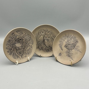 May include: Three small, round decorative plates with bird illustrations. Each plate features a different bird design in brown against a beige background. The plates are displayed on small metal stands, showcasing the detailed artwork.