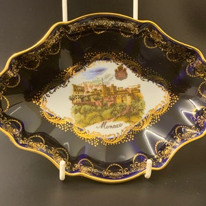 May include: A small, oval-shaped dish with a blue and gold design. The center of the dish features a white image of a building with a gold trim and the word "Monaco" printed below it.