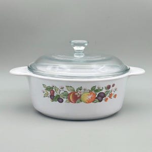 May include: White casserole dish with a glass lid. The dish features a colorful fruit design with peaches, plums, and berries. The dish has two handles and is suitable for cooking and serving.
