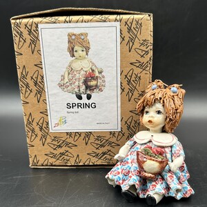 Può includere: Una statuina di bambola in ceramica di una ragazza con i capelli castani e un vestito floreale blu e bianco. Tiene un cestino di fiori ed è seduta su una superficie bianca. La bambola è etichettata "SPRING" e "Spring doll".