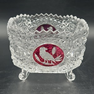 Könnte beinhalten: Eine klare, quadratische Kristallschale mit einem detaillierten, geometrischen Muster. Die Schale hat ein rotes Oval mit einem weißen Vogel- und Weizendesign. Die Schale hat kleine, abgerundete Füße und einen dekorativen Rand.