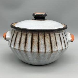 Può includere: Una pentola in ceramica con coperchio. La pentola è bianca con strisce verticali marroni e manici arancioni. Il coperchio è bianco con un pomello nero e arancione. La pentola è progettata per cucinare e servire.