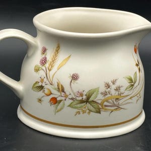 May include: Cream-coloured ceramic jug with a curved handle and spout. The jug is decorated with a floral design featuring berries, wheat, and leaves in shades of pink, green, and gold. A gold band accents the base.