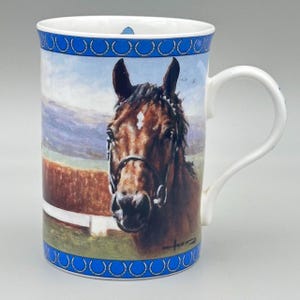 May include: White ceramic mug featuring a detailed painting of a brown horse's head with a white blaze. The mug has a blue horseshoe pattern around the top and bottom rims, and a white handle. The background shows a landscape with a fence and mountains.
