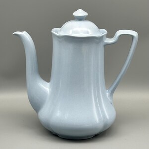 May include: A pale blue ceramic coffee pot with a lid and a curved handle. The pot has a spout and a rounded body. The lid has a small knob on top. The coffee pot is a light blue colour.