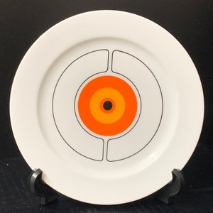 May include: White ceramic dinner plate with a black and white concentric circle design and a central orange, yellow, and black circle.