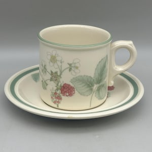May include: A cream-coloured ceramic cup and saucer set featuring a botanical illustration of green leaves, white flowers, and red raspberries. The rim of the cup and the edge of the saucer are accented with a thin, dark green circular band.