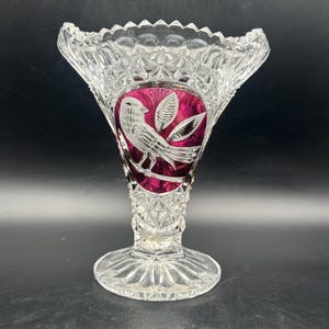 Può includere: Vaso di cristallo trasparente con parte superiore svasata e base rotonda. Il vaso presenta un design ovale rosso rubino con un motivo dettagliato di uccelli e foglie incise. Il cristallo è tagliato con un motivo a diamante.