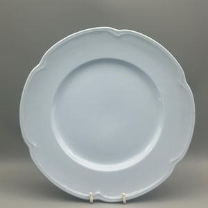 May include: A pale blue ceramic plate with a decorative, scalloped edge. The plate features a wide rim and a central recessed area. It is displayed on a white stand against a grey background. The plate is round.