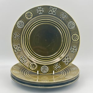 May include: A set of olive green ceramic plates featuring Celtic knot and geometric patterns. The plates are circular with a raised rim and a central recessed area. The plates are stacked, displaying the detailed designs. Each plate measures approximately 25 cm in diameter.