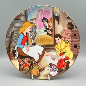 Può includere: Un piatto decorativo con una scena di Cenerentola. L'immagine raffigura Cenerentola, il Principe e vari personaggi animati. Il piatto ha un bordo dorato ed è ambientato su uno sfondo neutro.
