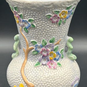 Könnte beinhalten: Eine hellblaue Keramikvase mit einer strukturierten Oberfläche und Blumendesigns. Die Vase hat zwei grüne Griffe und zeigt einen Zweig mit gelben und grünen Blättern und mehrfarbigen Blumen.