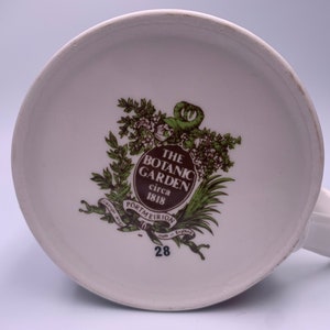 May include: The bottom of a white ceramic mug with a brown and green logo that reads "The Botanic Garden circa 1818 Portmeirion made in England" and the number 28.