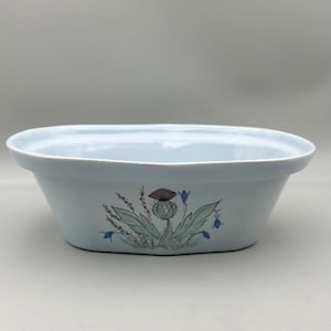 May include: A light blue ceramic serving dish with a thistle design. The oval dish has a light blue exterior and a decorative image of a thistle with green leaves, purple flower, and blue accents.