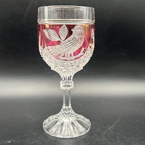 Könnte beinhalten: Ein klares Kristall-Weinglas mit rubinroter Innenseite. Das Glas hat ein geätztes Design eines Vogels, der auf einem Ast mit Blättern sitzt. Stiel und Fuß sind ebenfalls aus Kristall mit einem detaillierten Schliffmuster.