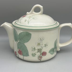 May include: A cream-coloured ceramic teapot featuring a botanical pattern of green leaves, white flowers, and red raspberries. The teapot has a matching lid with a loop handle, a curved spout, and a side handle, accented by a thin green rim.