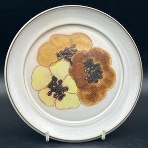 Può includere: Un piatto da portata in ceramica bianca con un bordo marrone e un motivo floreale con tre fiori gialli, arancioni e marroni con centri neri.