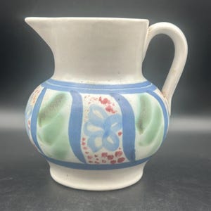 May include: A white ceramic pitcher with a curved handle and spout. The pitcher features a blue band with vertical stripes, and floral designs in blue, green, and red. The pitcher is a decorative item.