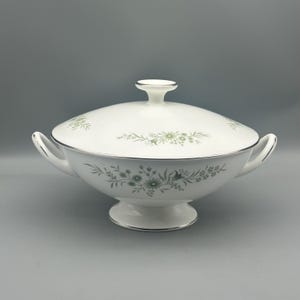 May include: White ceramic serving dish with lid, featuring a floral pattern in shades of green and gray. The dish has two handles and a pedestal base, with silver trim around the edges. A classic piece for serving food.