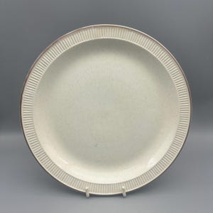 May include: A round, off-white ceramic plate with a speckled surface. The plate features a textured, ribbed border and a thin brown rim. This vintage plate is a classic piece of dinnerware.
