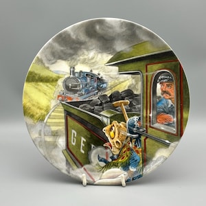 May include: Decorative plate featuring a whimsical train scene. A blue steam engine pulls a train along tracks, with a green train car in the foreground. The plate has a detailed illustration of a conductor and a cartoonish character.