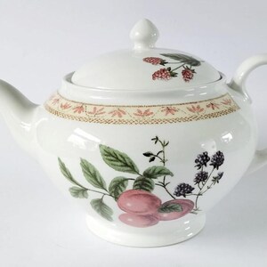Peut inclure: Théière en céramique blanche avec couvercle, bec et anse. La théière est ornée d'un motif floral avec des prunes roses, des feuilles vertes et des mûres. Une bande décorative entoure le pot près du haut.