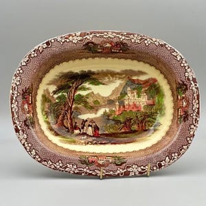 May include: Oval ceramic serving dish with a detailed landscape scene. The center features a castle, bridge, and figures near a tree. The border has a brown floral pattern. The dish is supported by a gold-colored metal stand.