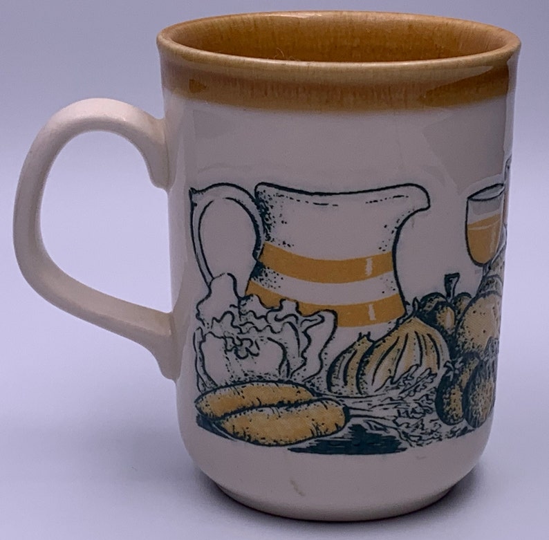 Puede incluir: Una taza de cer&aacute;mica blanca con un dise&ntilde;o amarillo y verde de una jarra, verduras y una copa de vino. La taza tiene un borde marr&oacute;n.