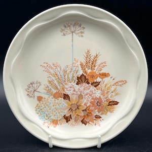 Peut inclure: Assiette en céramique crème avec un bord festonné et un motif floral. Le motif présente un bouquet de fleurs dans les tons orange, marron et jaune, avec des épis de blé et d'autres feuillages. L'assiette est soutenue par un support blanc.