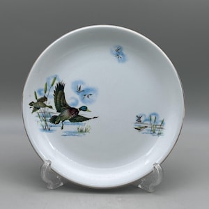 May include: A white ceramic plate featuring a colorful illustration of a mallard duck in flight over water with reeds. The design includes smaller flying ducks, a distant windmill, and a small boat, all finished with a thin silver metallic rim.