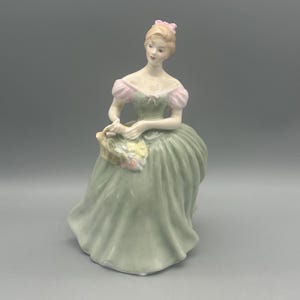 May include: A porcelain figurine of a seated woman in a long, flowing green dress with pink accents on the sleeves and a matching bow in her hair. She holds a small purse with floral details. The figurine has a classic, elegant design.