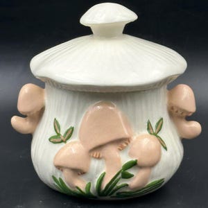 Può includere: Zuccheriera in ceramica bianca con coperchio a forma di fungo e design a fungo. La ciotola ha tre funghi rosa con foglie verdi sul davanti.
