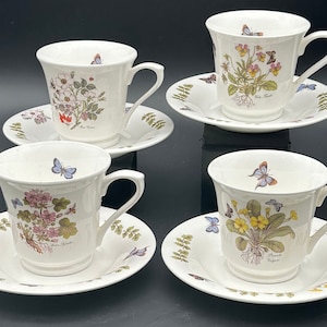 Könnte beinhalten: Set aus vier floralen Teetassen und Untertassen mit Schmetterlingsmotiven. Jede Tasse und Untertasse zeigt eine andere Blume und einen anderen Schmetterling. Die Tassen sind weiß mit einem Blumenmuster und goldenen Verzierungen. Die Untertassen sind weiß mit einem Blumenmuster.