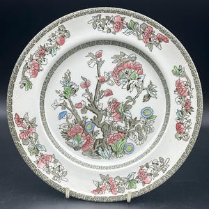 May include: A white ceramic plate with a floral design. The plate features a stylised tree with pink and green flowers and leaves. The border of the plate is decorated with a repeating geometric pattern in green and gold.