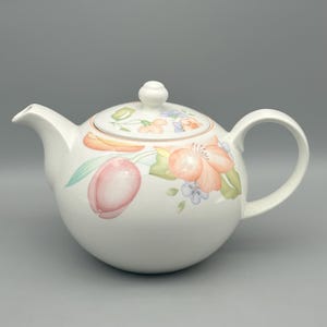 Può includere: Teiera in ceramica bianca con corpo arrotondato, beccuccio curvo e manico grande. La teiera è decorata con un motivo floreale con fiori rosa, arancioni e blu, insieme a foglie verdi. Il coperchio ha un piccolo pomello rotondo.