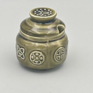 May include: A green ceramic jar with a lid and a spoon rest. The jar is adorned with embossed Celtic knot patterns. The lid features a decorative design. This jar is likely for storing condiments or spices, and is a decorative kitchen item.