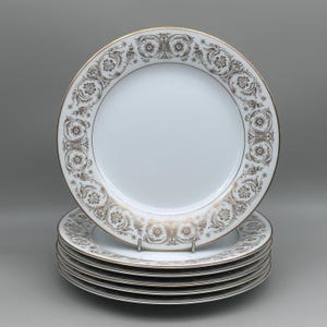 May include: A stack of six white porcelain plates featuring a wide rim decorated with a metallic gold and brown floral scrollwork pattern. Each plate has a thin gold-toned trim along the outer edge, creating a uniform and cohesive set of dinnerware.