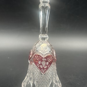 May include: Clear glass bell with a heart-shaped handle and a heart design on the bell. The heart design is red with a floral pattern in the center. The bell has a diamond-cut pattern on the lower portion.