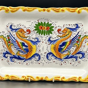 May include: A white ceramic serving tray with two yellow, blue and red dragons on either side of a green floral design. The tray has two yellow handles.