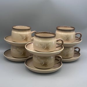 May include: A set of four ceramic cups and saucers in a light beige color with a darker brown rim and a floral pattern on the front. Each cup features a side handle and sits on a matching round saucer. The set is arranged in a stacked formation.