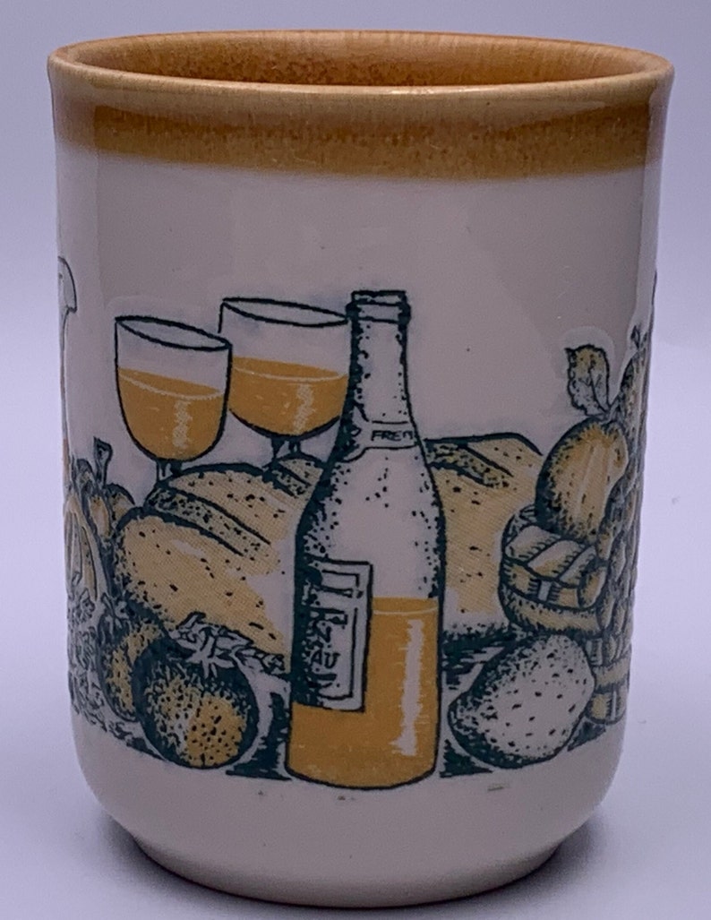 Puede incluir: Una taza de cer&aacute;mica blanca con un borde amarillo y un dise&ntilde;o verde y amarillo de una botella de vino, dos vasos de vino, pan, fruta y fresas.