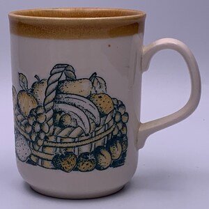 Puede incluir: Una taza de cer&aacute;mica blanca con un borde marr&oacute;n y un dise&ntilde;o de cesta de frutas. La cesta est&aacute; llena de manzanas, peras, pl&aacute;tanos, uvas y fresas. La fruta est&aacute; dibujada en negro y verde con acentos amarillos.