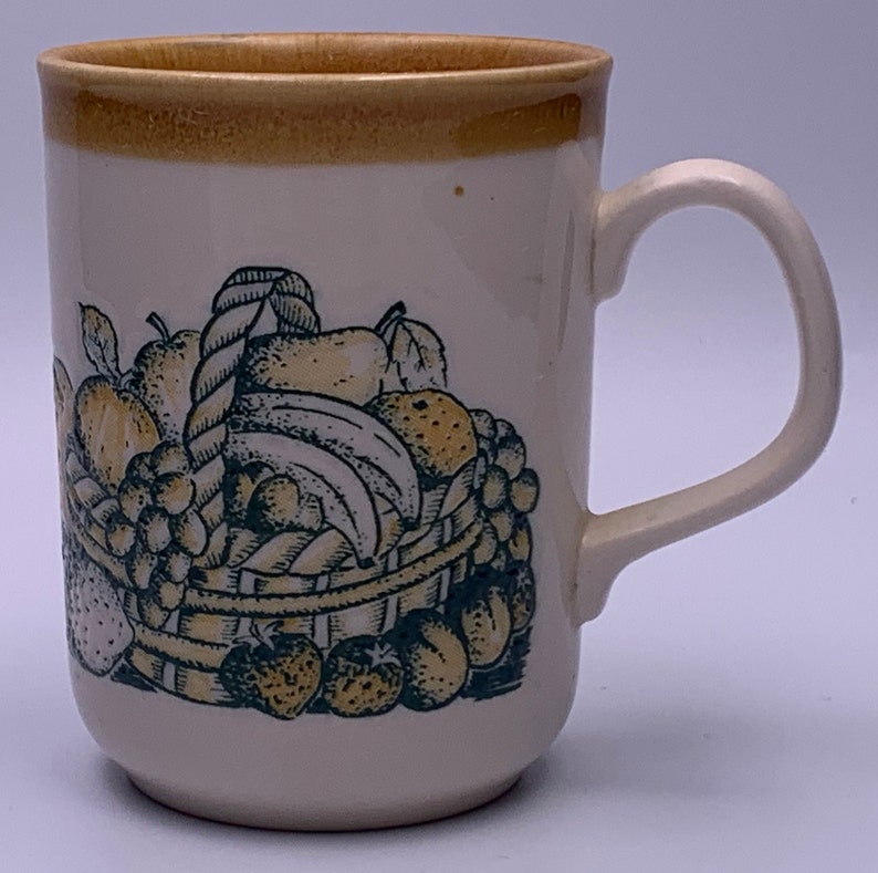 Puede incluir: Una taza de cer&aacute;mica blanca con un borde marr&oacute;n y un dise&ntilde;o de cesta de frutas. La cesta est&aacute; llena de manzanas, peras, pl&aacute;tanos, uvas y fresas. La fruta est&aacute; dibujada en negro y verde con acentos amarillos.