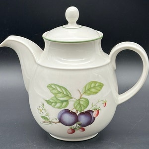 Può includere: Teiera in ceramica bianca con un bordo verde e un design floreale con prugne e fragole. La teiera ha un coperchio e un manico.