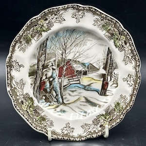 May include: A vintage ceramic plate depicting a winter scene. The plate has a central image of a red barn in a snowy landscape, with a brown and green floral border. The plate has a scalloped edge.