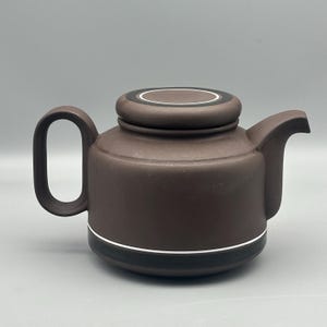 Peut inclure: Une théière en céramique marron avec un corps arrondi, une anse incurvée et un bec verseur. Le couvercle est plat avec une bordure noire et blanche. Une bande noire et blanche entoure la base de la théière. La théière est conçue pour infuser du thé.