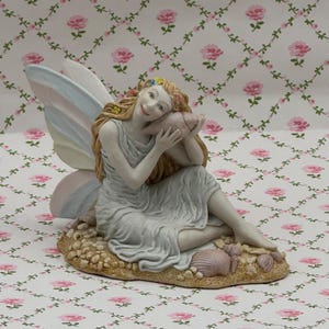 May include: A fairy figurine, seated on a sandy base with seashells. The fairy wears a flowing grey dress and holds a seashell to her ear. Her long, flowing hair is adorned with flowers, and she has pastel-coloured wings.
