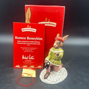 Può includere: Una statuina Royal Doulton Bunnykins di Romeo, in ginocchio su una base a forma di nuvola. La statuina indossa una giacca verde, un cappello rosso e una sciarpa viola. La scatola e le carte sono visibili, con il testo "Romeo Bunnykins".