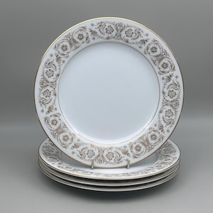May include: A stack of white ceramic plates featuring a wide rim decorated with a brown floral scroll pattern and a thin gold trim along the outer edge. The center of each plate is plain white, and the design repeats around the entire border.